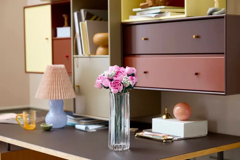 LEGO Bouquet of Pink Roses in a vase on a dark desk, surrounded by colorful cabinets and decor.