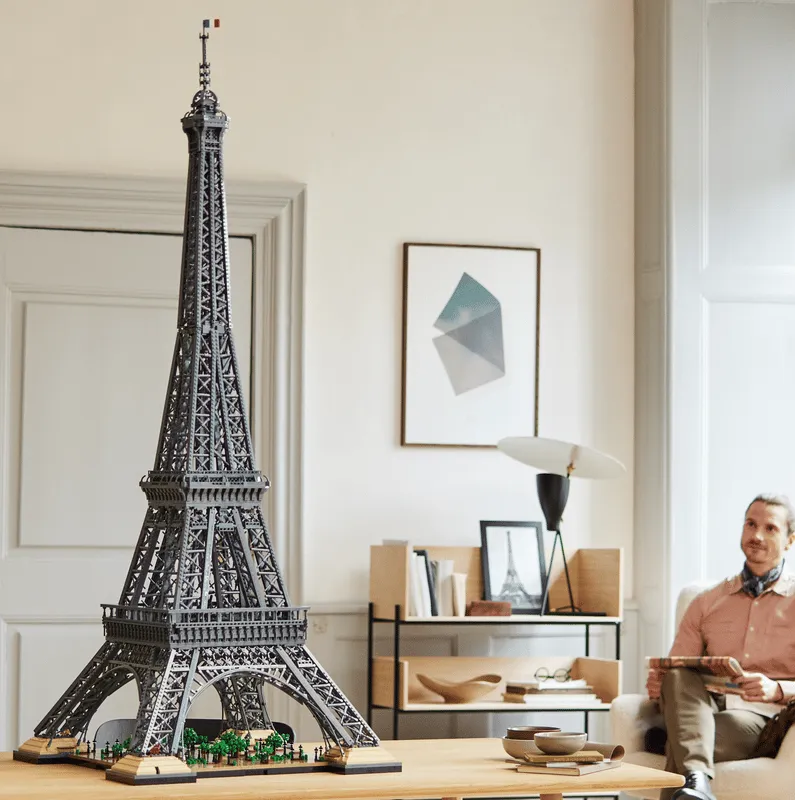 A man sitting on a couch in the background, looking at the impressive completed LEGO Eiffel Tower 10307 model displayed prominently on a wooden table in a well-decorated room.