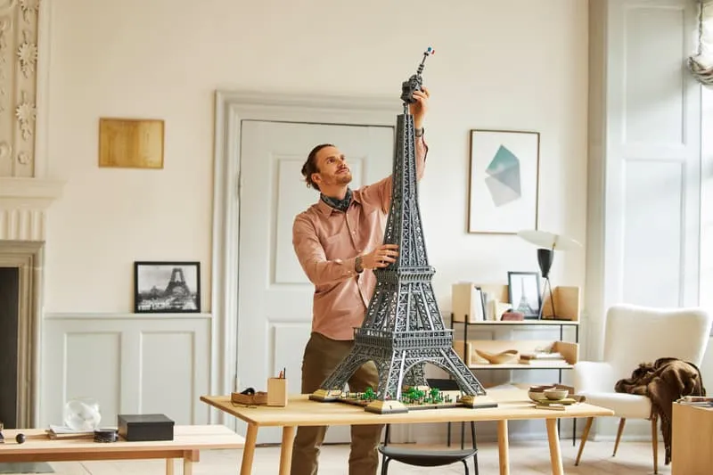 A man in a light-colored shirt is standing, attaching the topmost section of the LEGO Eiffel Tower 10307 model, which is placed on a wooden table in a well-lit room.