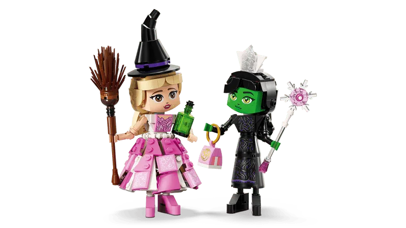Lego Elphaba and Glinda figures swapped accessories: Glinda wearing the witch's hat and holding a potion, while Elphaba wears Glinda's crown and holds the wand, against a white background.