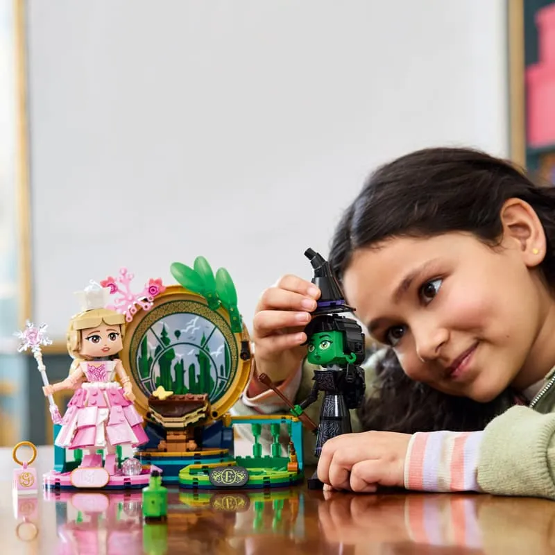 A young girl smiling and playing with the Lego Elphaba & Glinda figures, adjusting Elphaba's witch hat, on a wooden table.