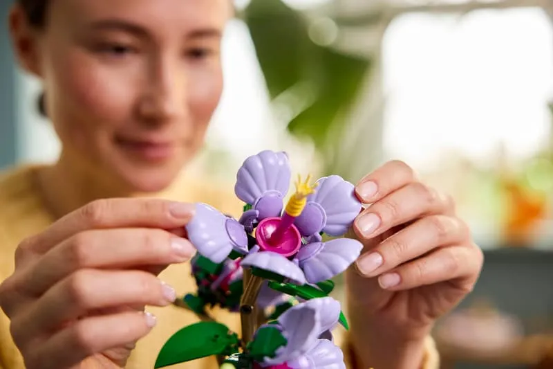 A person's hands gently adjusting one of the Lego Hibiscus flowers, with a blurred background suggesting a home environment.