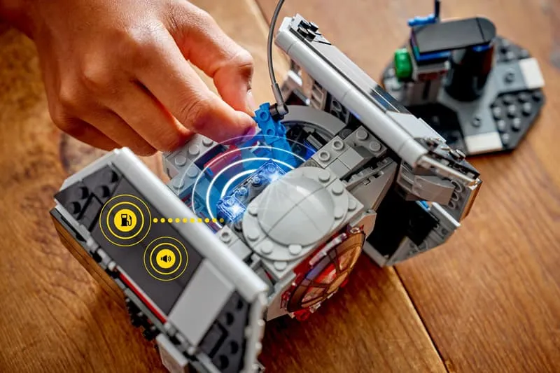 A close-up of Darth Vader's TIE Fighter connected to a small black base unit via a blue cable, with a glowing light effect emanating from the connection point, indicating power or data transfer.