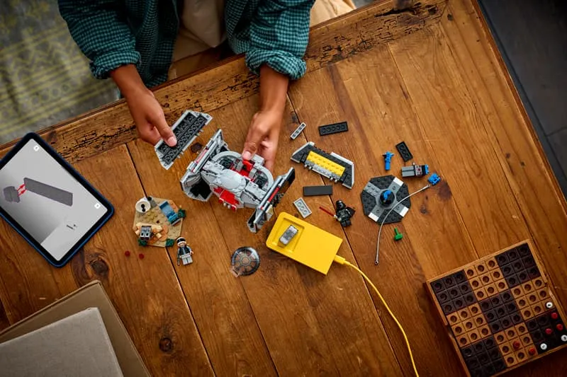 An overhead view of a child's hands playing with the LEGO Star Wars 75421 set on a wooden table, showing the TIE Fighter, Rebel turret, minifigures, Smart Charger, and a tablet displaying digital building instructions.