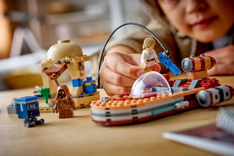 A child's hands playing with the LEGO Star Wars: Luke's Landspeeder 75420 set, specifically connecting a part to the X-34 Landspeeder with Luke Skywalker and the Jawa minifigures nearby, and the moisture vaporator in the background.