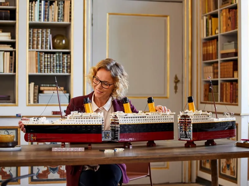 Lifestyle shot of a woman in glasses assembling the three modular sections of the LEGO Titanic model on a wooden table in a room with bookshelves.