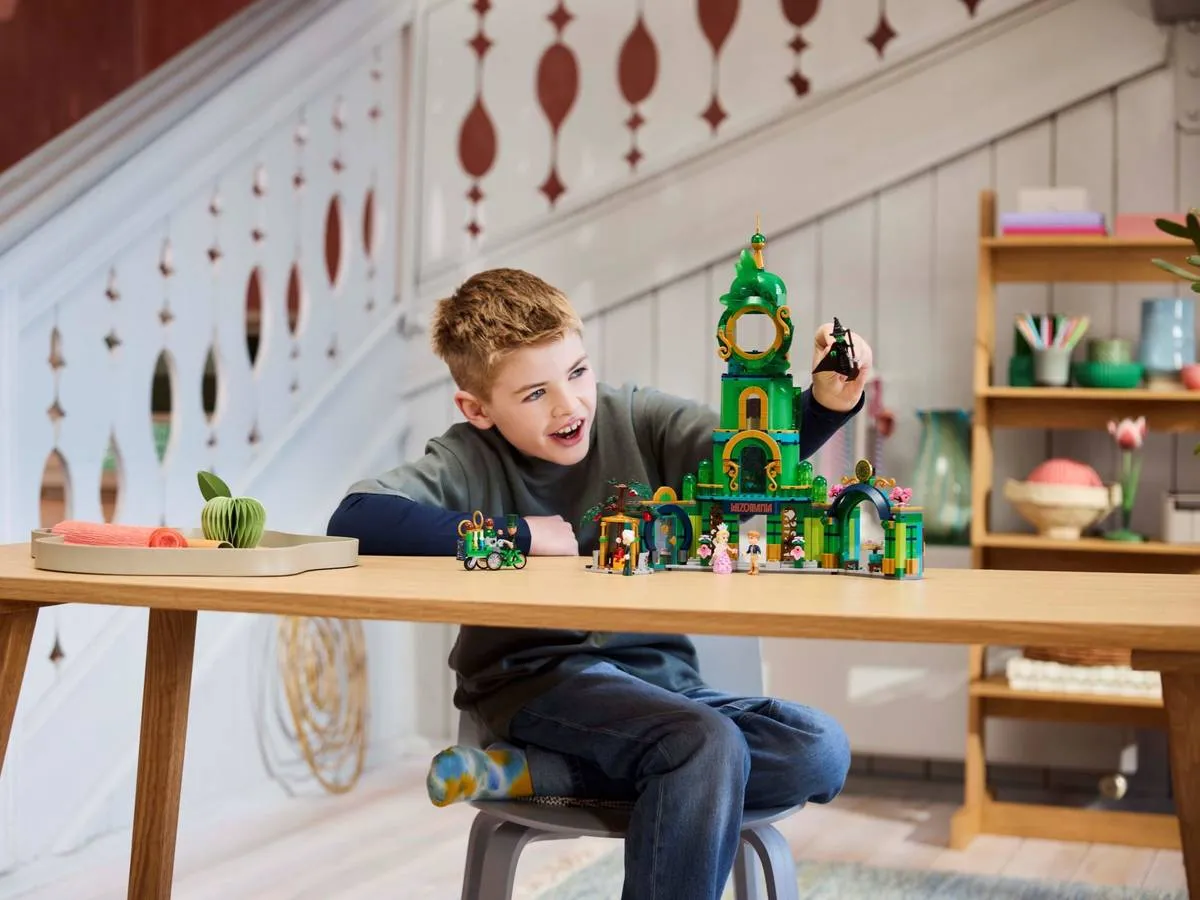 A young boy, smiling enthusiastically, playing with the Lego Welcome to Emerald City 75684 set on a wooden table in a brightly lit room with stairs in the background.