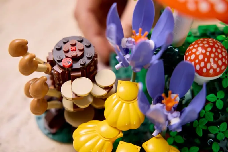 A hand gently inserting a piece into the LEGO Woodland Mushrooms set, specifically near the brown tree stump with tan mushrooms, yellow shell-shaped fungi, and purple flowers.